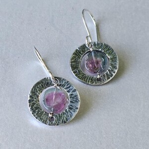 Peut inclure: Une paire de boucles d'oreilles pendantes avec un cadre circulaire argenté à la finition martelée. Chaque boucle contient une pierre précieuse ronde, translucide, avec un dégradé rose et vert, suspendue par un crochet en métal argenté.