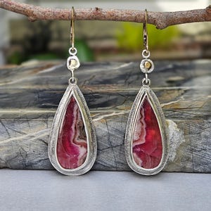 Puede incluir: Un par de pendientes en forma de lágrima con incrustaciones de piedra pulida en bandas de color rosa oscuro y blanco, engastados en marcos de plata texturizada. Cada pendiente cuelga de un gancho dorado con un pequeño acento circular.