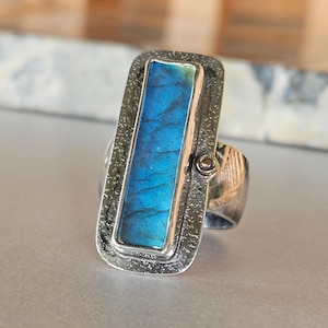 May include: A rectangular silver ring featuring a central elongated labradorite stone with a vibrant blue iridescent flash. The metal setting has a dark, textured, hammered finish and includes a small, circular accent stone on the right side.
