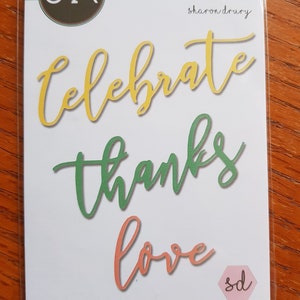 May include: A white card with the words "celebrate", "thanks", and "love" written in yellow, green, and orange cursive lettering. The words are cut out of the card and are ready to be used for crafting.