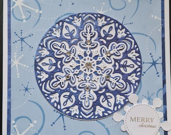 May include: A festive Christmas card with a detailed blue and white snowflake design, embellished with rhinestones. The card has a light blue backdrop with snowflake and swirl motifs, and a small tag that reads "MERRY Christmas".