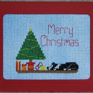 May include: A cross-stitch card with a green Christmas tree, presents, and a black and white dog. The card has a red border and says "Merry Christmas" in red.
