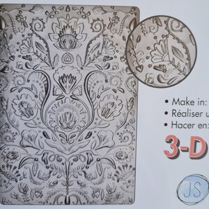 May include: A grey 3D embossing plate featuring an intricate floral pattern. The design includes flowers, leaves, and swirling motifs. A circular inset provides a close-up view of the pattern. Text on the right side says "Make in: Réaliser un Hacer en: 3-D".