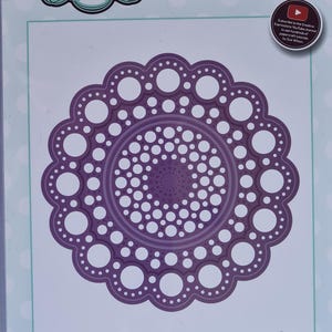 May include: A purple steel craft die set with a circular, floral design. The die has a diameter of 10.4 cm and is labeled "FRAMES & TAGS - ARIEL". The packaging includes the text "CREATIVE EXPRESSIONS" and "Craft Dies by Sue Wilson".