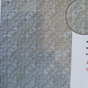 May include: A textured, grey surface with a repeating floral pattern. The design features a series of four-petal flowers connected by curved lines, creating a grid-like arrangement. A circular inset shows a close-up of the pattern.