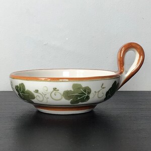 May include: A small white ceramic bowl with a brown rim and a green vine design. The bowl has a single handle on the side.