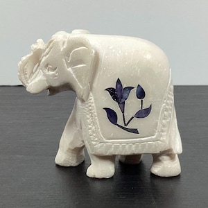 May include: A small, off-white elephant figurine with a floral design in dark blue on its back. The elephant has detailed carvings and is standing on four legs. The figurine is made of a solid material and has a smooth texture.