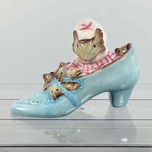 以下が含まれることがあります： ハイヒール付きのライトブルーのセラミックシューズの置物。リボンと小さなネズミのフィギュアで飾られています。靴の中には、ピンクと白のチェック柄のドレスと白いボンネットを着たネズミが入っています。