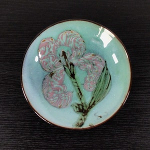 May include: A small, round, blue ceramic plate with a pink and green floral design. The plate has a brown rim.