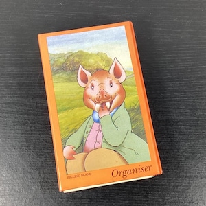 May include: An orange organiser book with a cartoon pig character in a green coat and pink shirt. The book has the word "Organiser" printed on the bottom right and "Pigling Bland" on the bottom left.