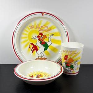 May include: A breakfast set with a plate, bowl, and cup, all decorated with a rooster design. The plate and cup feature the text "The Sunshine Breakfast". The plate has a red and green border, and the bowl has a red border.