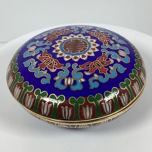 Könnte beinhalten: Eine runde, blaue Cloisonné-Box mit goldenem Rand. Der Deckel hat ein zentrales Design mit roten, weißen und goldenen Akzenten, umgeben von einem floralen Muster. Die Box hat einen Durchmesser von etwa 7,6 cm.