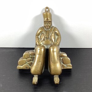 May include: A brass figurine of a person wearing a tall hat and jacket, sitting on a stone wall. The figure has a loop at the top for hanging and features a metallic gold finish with visible details on the clothing and the textured stone base.