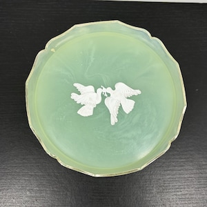 May include: A round, light green, translucent tray with a scalloped edge and a metallic silver rim. The centre features a white, raised relief design of two birds facing each other, with one bird holding a small sprig in its beak.