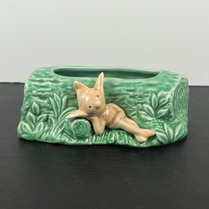 May include: A green ceramic planter shaped like a log, with a reclining figurine of a nude bunny. The planter has a rectangular opening and is decorated with embossed foliage. The bunny is light brown.