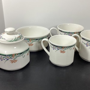 May include: A white ceramic tea set with floral accents. The set includes a sugar bowl with a lid, a serving bowl, and three mugs. The floral design features orange, blue, and green flowers with a black and green border.