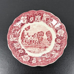 May include: A round, red and white decorative plate with a scalloped edge. The plate features a central landscape scene with a building, trees, and a boat on a body of water. Floral patterns surround the central image.