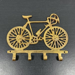 May include: A gold-toned metal wall hook rack shaped like a bicycle. The design features two large spoked wheels, a frame, handlebars, and a seat. At the base, there are four downward-facing hooks for hanging items, with two small screw holes.