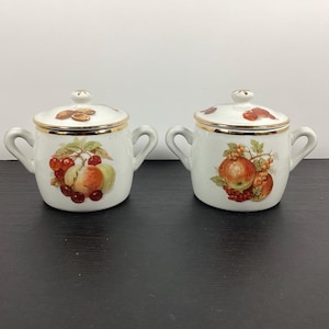 May include: Two white ceramic sugar bowls with lids and side handles. Each bowl features a gold-trimmed rim and a colourful illustration of assorted fruits, including cherries, peaches, and grapes, on the front and on the top of the lid.