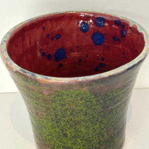 May include: A ceramic cup with a red interior and green exterior. The interior has blue speckles. The exterior has a mottled green glaze.