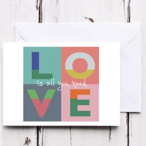 May include: A greeting card with the word "LOVE" in colorful block letters. The text "is all you need" is written in cursive below the "L" and "O". The card is inside a white envelope, set against a white wooden background.
