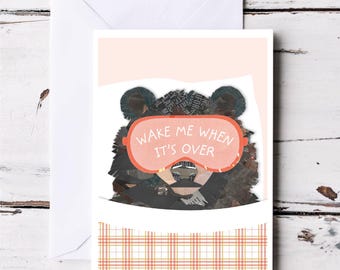 May include: A greeting card featuring a black bear wearing pink sleep mask with the text "Wake me when it's over". The card has a pink background and a plaid pattern at the bottom.