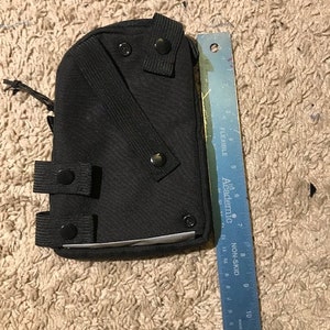 K&ouml;nnte beinhalten: Schwarze Tasche mit mehreren Riemen und Druckkn&ouml;pfen. Ein blaues Lineal mit Zentimetermarkierungen liegt neben der Tasche. Auf dem Lineal steht "FLEXIBLE" und "NON-SKID".