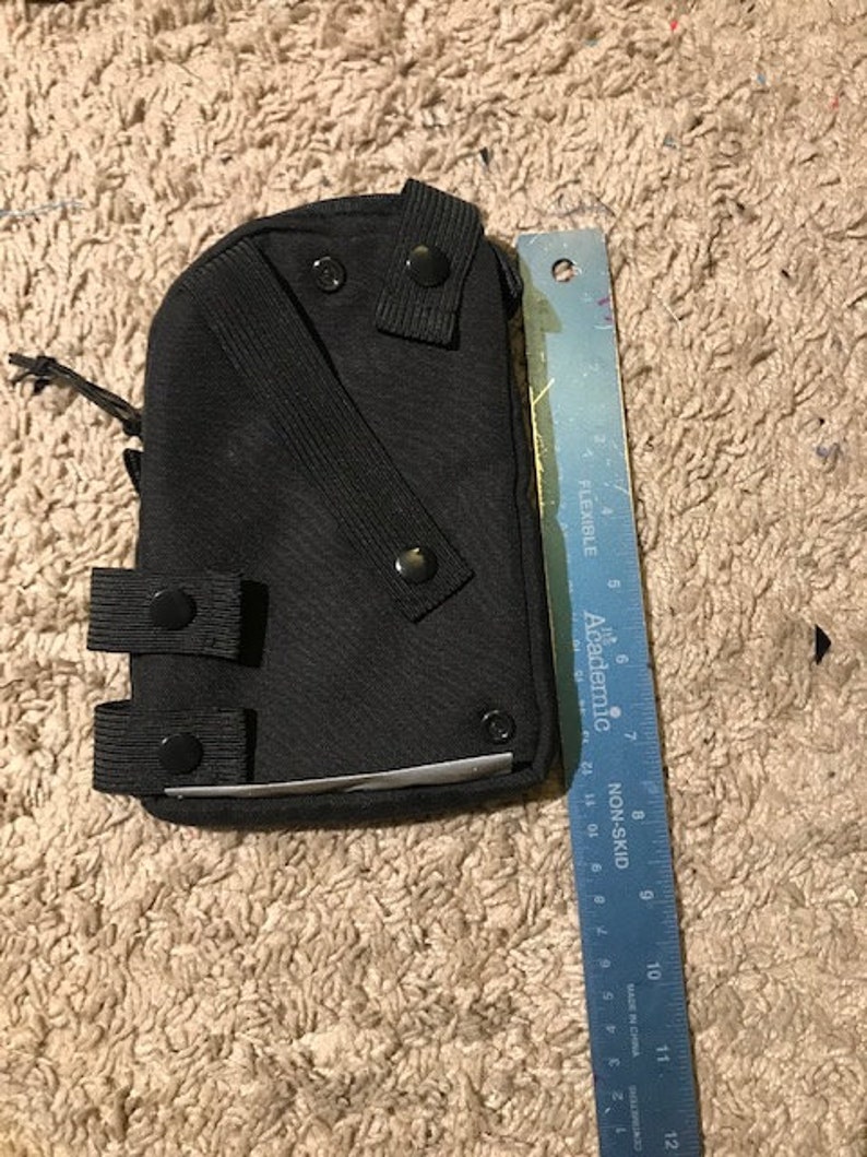 K&ouml;nnte beinhalten: Schwarze Tasche mit mehreren Riemen und Druckkn&ouml;pfen. Ein blaues Lineal mit Zentimetermarkierungen liegt neben der Tasche. Auf dem Lineal steht "FLEXIBLE" und "NON-SKID".