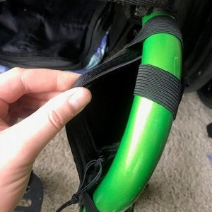 K&ouml;nnte beinhalten: Nahaufnahme einer schwarzen Tasche mit Rei&szlig;verschluss, befestigt an einer hellgr&uuml;nen, gebogenen Metallstange. Die Stange ist mit schwarzen Gummib&auml;ndern umwickelt. Die Tasche ist ge&ouml;ffnet und zeigt ein schwarzes Innenleben.