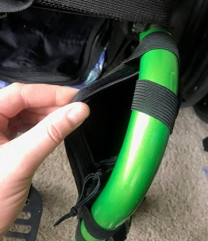 K&ouml;nnte beinhalten: Nahaufnahme einer schwarzen Tasche mit Rei&szlig;verschluss, befestigt an einer hellgr&uuml;nen, gebogenen Metallstange. Die Stange ist mit schwarzen Gummib&auml;ndern umwickelt. Die Tasche ist ge&ouml;ffnet und zeigt ein schwarzes Innenleben.