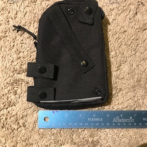 K&ouml;nnte beinhalten: Eine schwarze Tasche mit mehreren Knopfverschl&uuml;ssen und einem Riemen. Die Tasche hat eine rechteckige Form und ist etwa 15 cm lang, gemessen mit dem blauen Lineal im Bild. Das Lineal tr&auml;gt die Aufschrift "FLEXIBLE Academic".