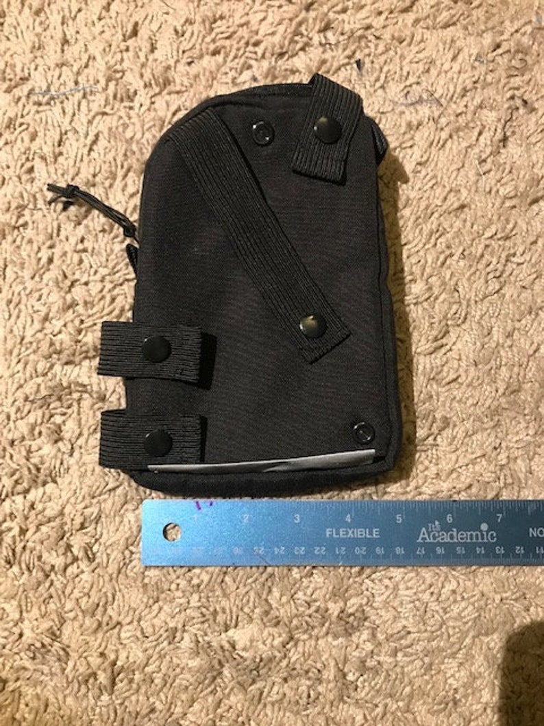 K&ouml;nnte beinhalten: Eine schwarze Tasche mit mehreren Knopfverschl&uuml;ssen und einem Riemen. Die Tasche hat eine rechteckige Form und ist etwa 15 cm lang, gemessen mit dem blauen Lineal im Bild. Das Lineal tr&auml;gt die Aufschrift "FLEXIBLE Academic".