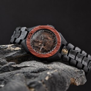 May include: A wristwatch featuring a dark black wooden link band and a circular reddish-brown wooden bezel. The watch face displays a detailed relief of a character and the word Fallout. A small gold-coloured lightning bolt emblem is on the band.