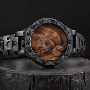 May include: A wristwatch featuring a dark black wooden band and a matching black wooden bezel with intricate carved details. The watch face displays a natural brown wood grain background with a three-dimensional carved relief of a human figure.