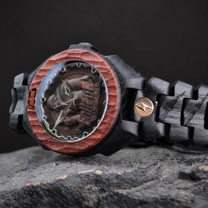 May include: A wristwatch with a black wooden link band and a carved reddish-brown wooden bezel. The watch face features a Fallout New Vegas graphic with the text Fallout and New Vegas visible. A small circular gold-toned emblem is on the band.