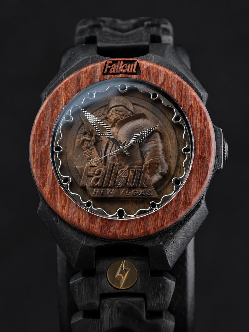May include: A wristwatch with a black frame and a reddish-brown wooden bezel. The watch face features a relief of a character in armour with the text Fallout and New Vegas. The band is black with a small circular gold-toned emblem featuring a lightning bolt.