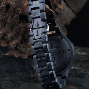 May include: Close-up of a black wooden wristwatch with a matching band. The watch face and band are made of dark wood, with the brand name "STAD" visible on the watch face. The band features unique carved details.
