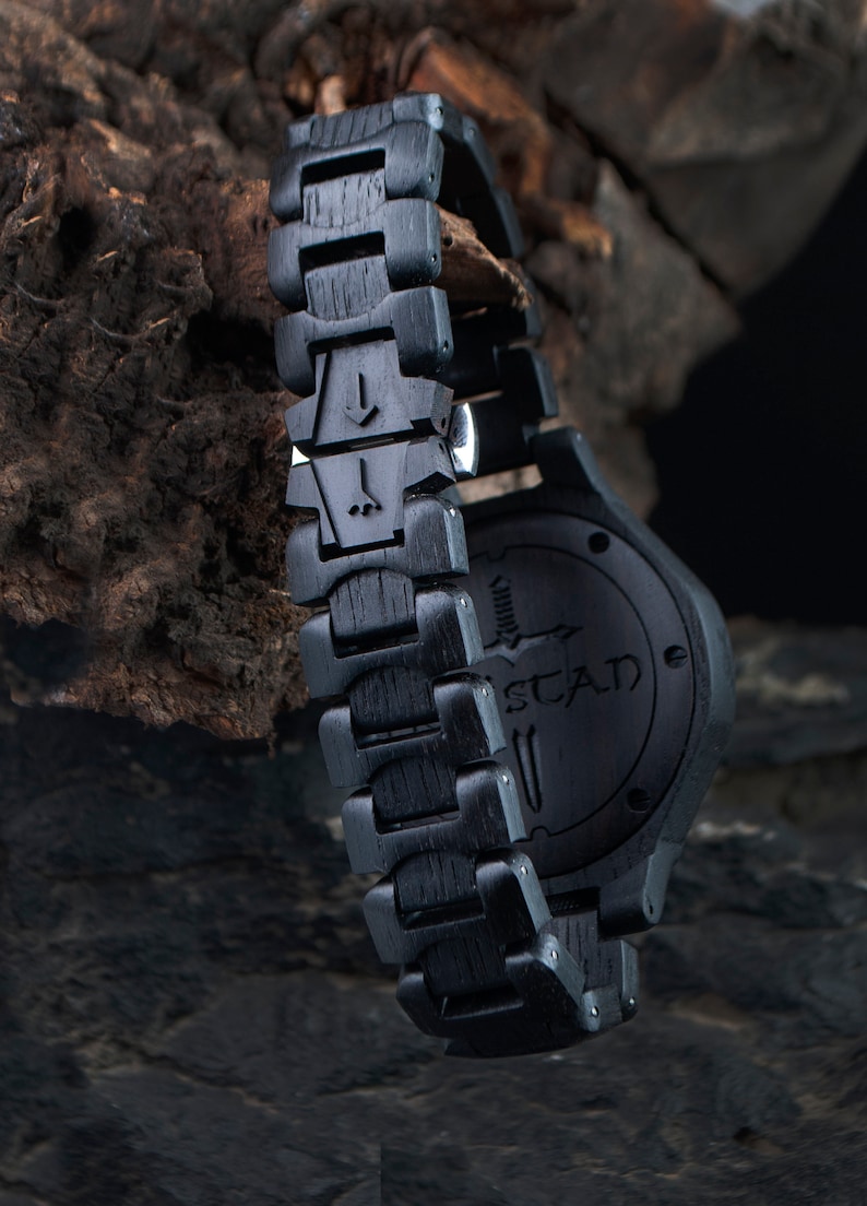 May include: Close-up of a black wooden wristwatch with a matching band. The watch face and band are made of dark wood, with the brand name "STAD" visible on the watch face. The band features unique carved details.