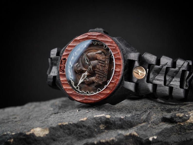May include: A wristwatch featuring a dark black wooden band and a textured reddish-brown wooden bezel. The watch face displays the Fallout New Vegas logo with metallic hands. A small gold-toned circular accent is visible on the watch link.