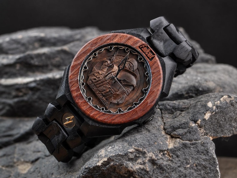 May include: A wooden wristwatch featuring a dark brown circular bezel and a black wooden link band. The watch face displays a Fallout New Vegas graphic with metallic hands and the Fallout logo. A small gold lightning bolt emblem is on the band.