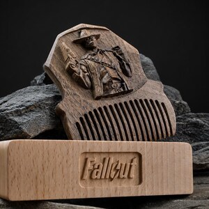 May include: A dark brown wooden comb featuring a carved relief of a figure wearing a hat and holding a firearm. In front of the comb sits a light-coloured rectangular wooden block with the word Fallout embossed in the centre of the front face.