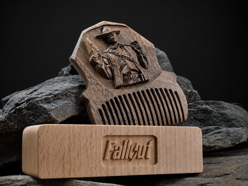 May include: A dark brown wooden comb featuring a carved relief of a figure wearing a hat and holding a firearm. In front of the comb sits a light-coloured rectangular wooden block with the word Fallout embossed in the centre of the front face.