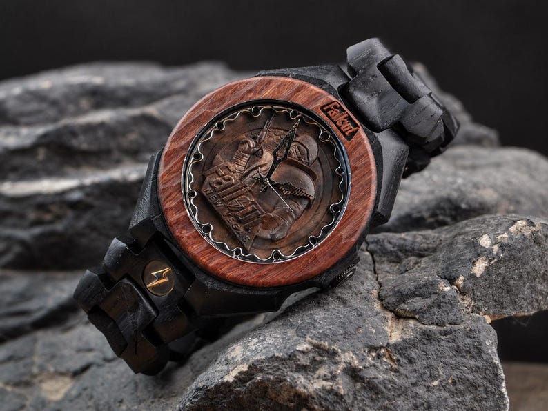 May include: A wooden wristwatch featuring a dark brown circular bezel and a black wooden link band. The watch face displays a Fallout New Vegas graphic with metallic hands and the Fallout logo. A small gold lightning bolt emblem is on the band.