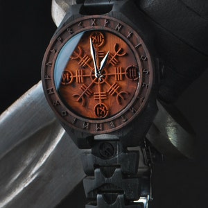 Puede incluir: Un reloj de pulsera de madera con una banda negra y una cara circular con un fondo de madera roja con un diseño de brújula de plata y runas alrededor del borde.