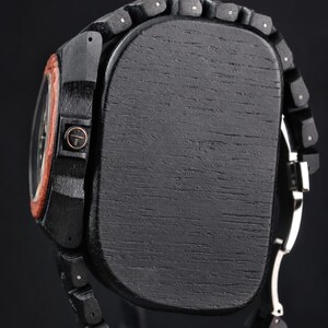 May include: A wristwatch featuring a black wooden case and a matching black wooden link band. The side of the case displays a circular crown with a cross symbol. A reddish-brown wooden bezel surrounds the watch face, and a silver metal clasp is visible.