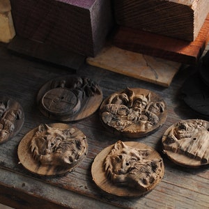 May include: A collection of intricately carved wooden coasters. The coasters feature detailed relief carvings of Viking figures, wolves, and other designs. The wood has a natural, warm brown tone, and the carvings are highlighted by the shadows.