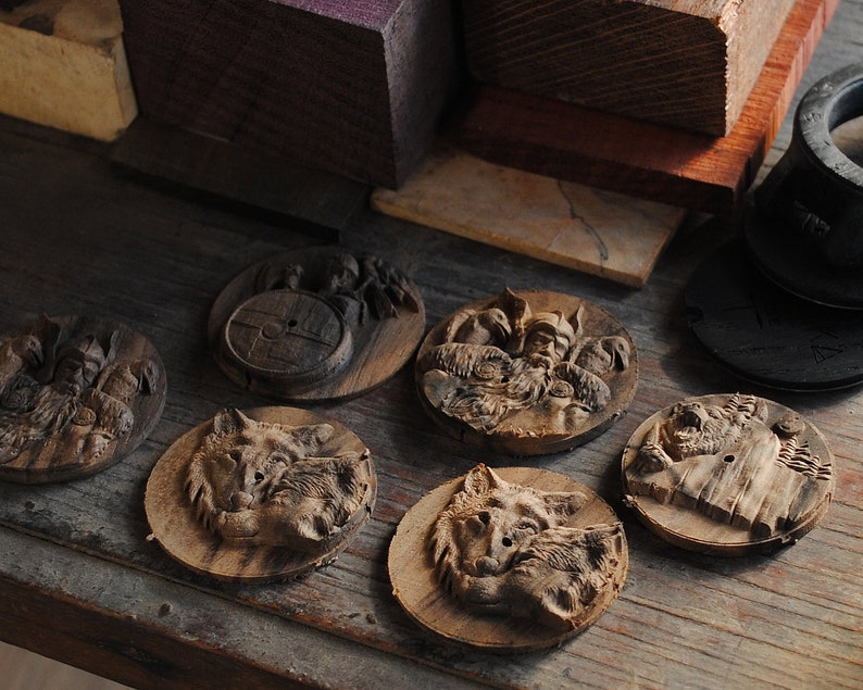 May include: A collection of intricately carved wooden coasters. The coasters feature detailed relief carvings of Viking figures, wolves, and other designs. The wood has a natural, warm brown tone, and the carvings are highlighted by the shadows.