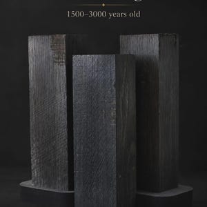 May include: Three rectangular blocks of dark, textured bog oak arranged vertically. The text above reads Immortal Bog Oak 1500-3000 years old. The wood displays a deep charcoal color with visible grain patterns and a weathered, aged surface texture.