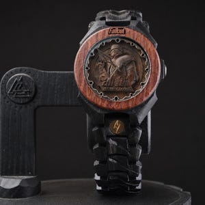 May include: A black wooden wristwatch featuring a circular face with a brown wood bezel. The dial displays a relief of a character in armour with the text Fallout and Fallout New Vegas. The band is black wood with a small gold lightning bolt emblem.