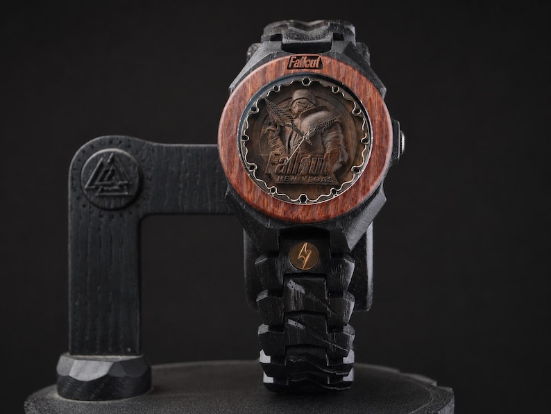 May include: A black wooden wristwatch featuring a circular face with a brown wood bezel. The dial displays a relief of a character in armour with the text Fallout and Fallout New Vegas. The band is black wood with a small gold lightning bolt emblem.